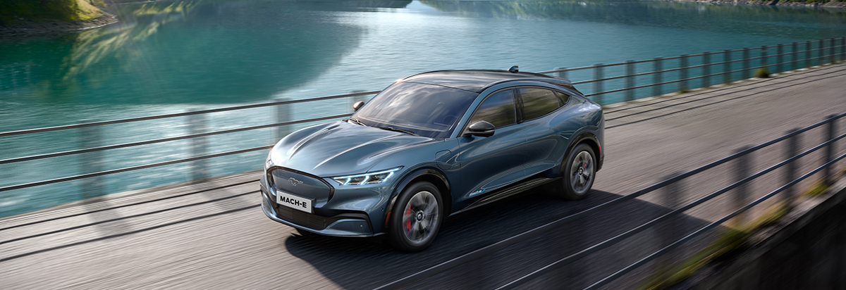 Ein blauer Ford Mustang Mach-E fährt auf einer Straße neben einem ruhigen See.